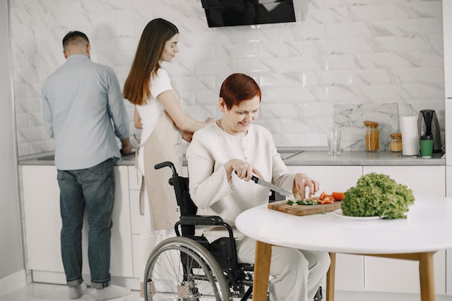 A family prepares dinner in a modern kitchen as the children wash dishes while their parent in a wheelchair chops vegetables on the table.